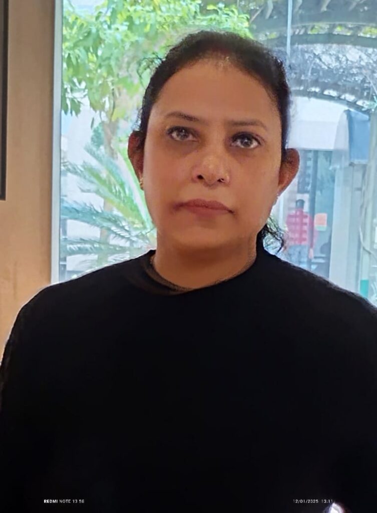 A woman with dark hair, wearing a black top, stands in front of a window with greenery visible outside.