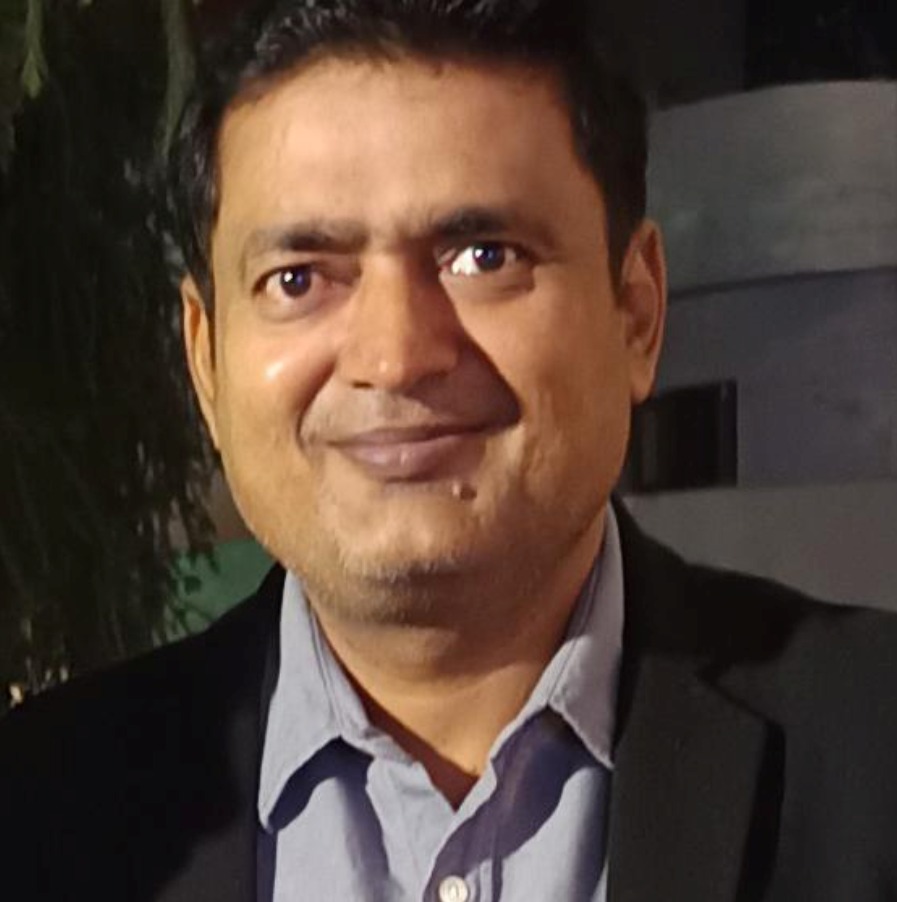 A man smiling in a dark suit with a light shirt, standing outdoors with greenery in the background.