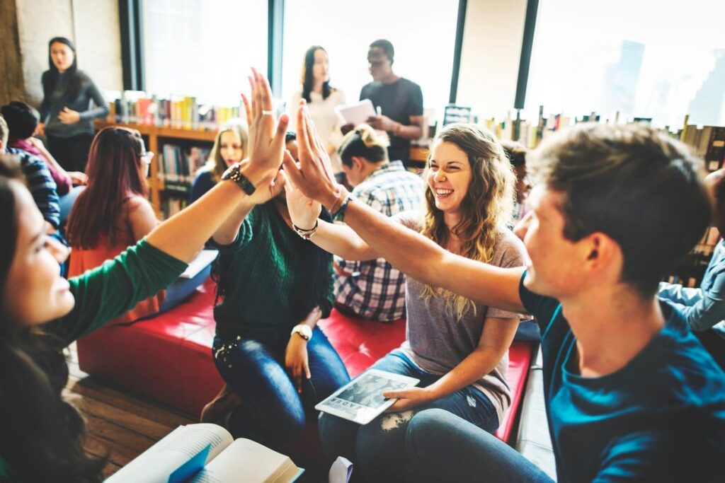 A group of young people are smiling and interacting in a lively setting, likely a library or study space.